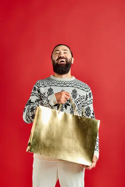 Lächelnder bärtiger Mann im Pullover mit Ornament, der Einkaufstüten vor rotem Hintergrund hält, Weihnachten — Stockfoto