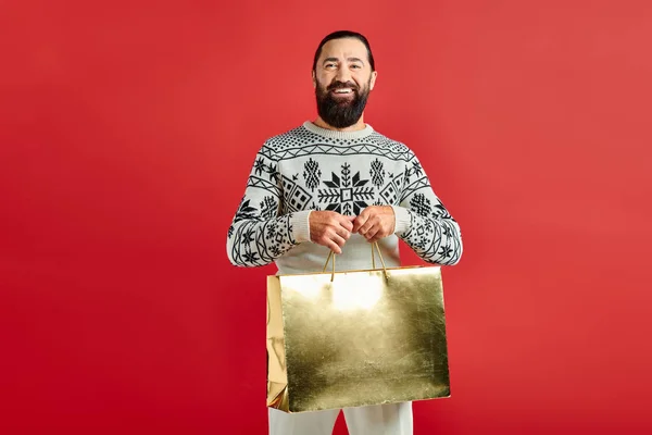 Sonriente hombre barbudo en suéter con adorno sosteniendo bolsas de compras sobre fondo rojo, Feliz Navidad - foto de stock