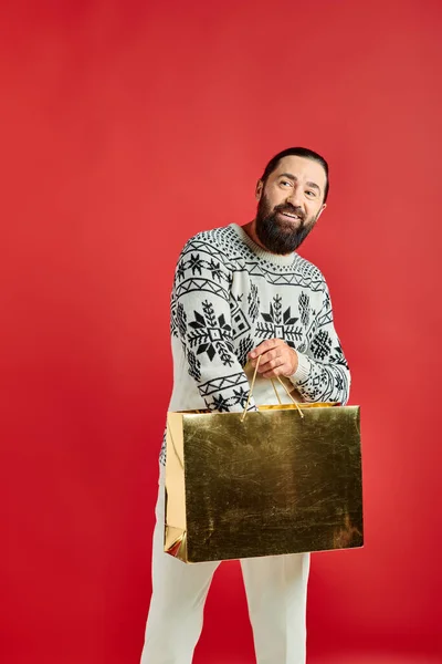 Весёлый бородатый мужчина в рождественском свитере держит сумку с покупками на красном фоне, праздничные распродажи — стоковое фото