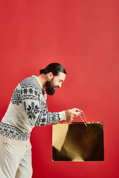 Весёлый бородатый мужчина в рождественском свитере смотрит внутрь сумки с покупками на красном фоне, продажи — стоковое фото