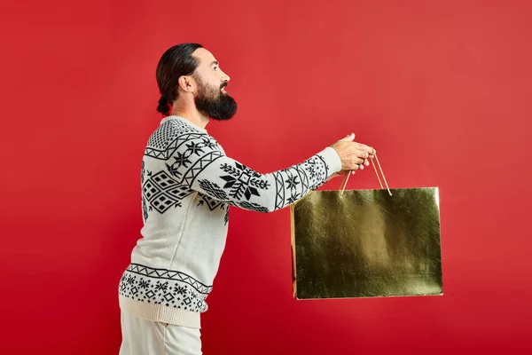 Homme barbu gai en pull de Noël tenant sac à provisions sur fond rouge, les ventes de vacances — Photo de stock