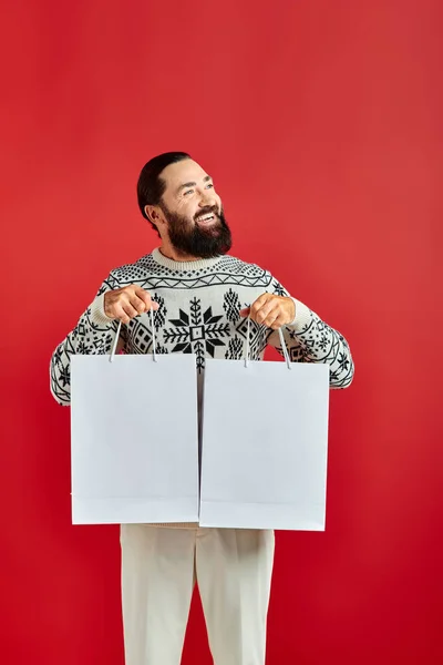 Homme barbu positif en pull de Noël tenant des sacs à provisions sur fond rouge, les ventes de vacances — Photo de stock
