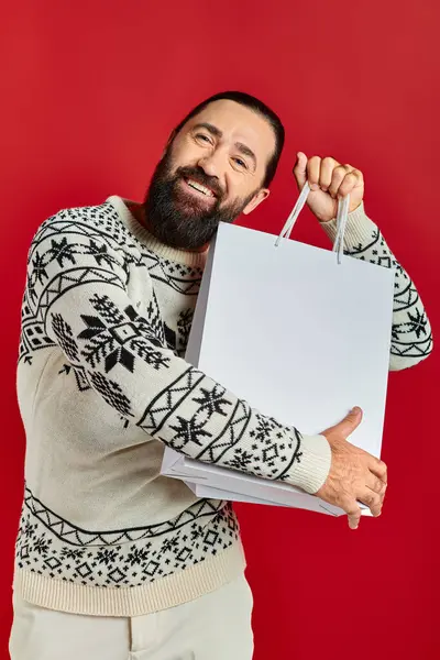 Barbudo hombre feliz en suéter de Navidad con bolsas de compras en el fondo rojo, las ventas de vacaciones - foto de stock