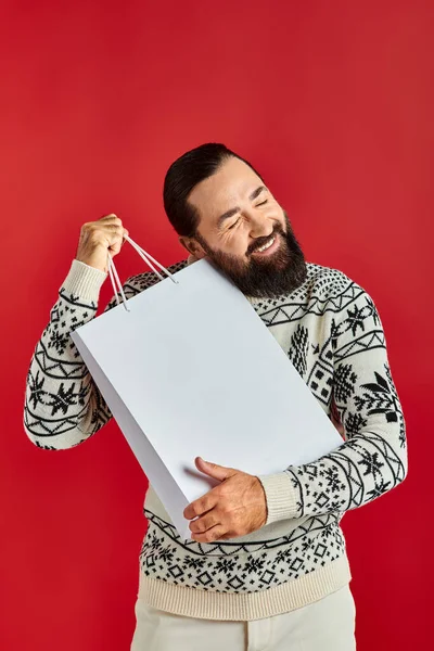 Весёлый бородатый мужчина в рождественском свитере держит сумки с покупками на красном фоне, праздничные распродажи — стоковое фото
