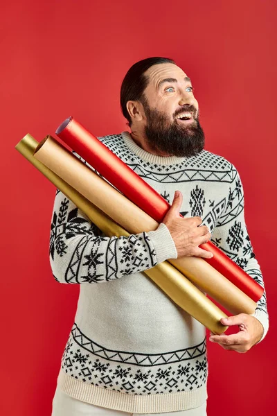 Thrilled bearded man in Christmas sweater holding wrapping paper on red backdrop, holiday sales — Stock Photo