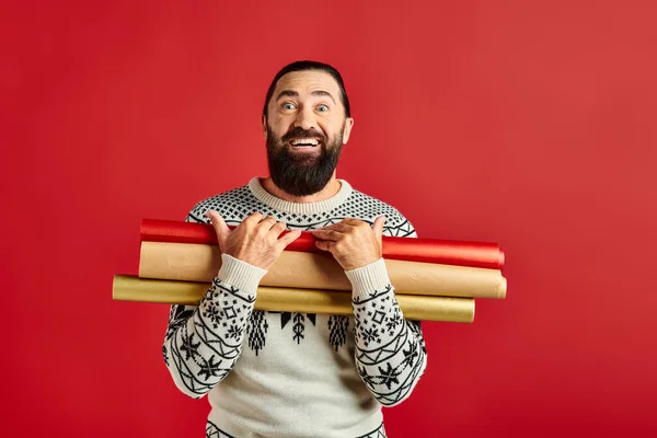 Счастливый бородатый мужчина в рождественском свитере держа оберточную бумагу на красном фоне, праздничные продажи — стоковое фото
