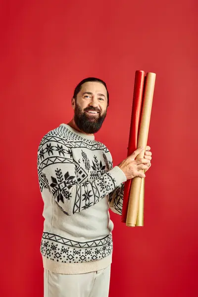 Hombre barbudo alegre en suéter de invierno celebración de papel de regalo de colores sobre fondo rojo, Feliz Navidad - foto de stock