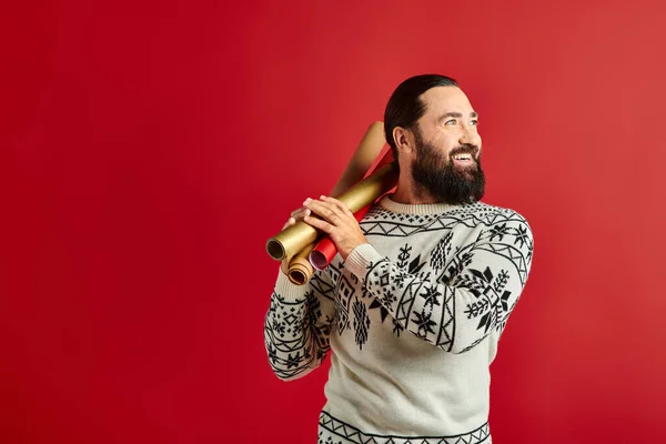 Веселый бородатый мужчина в зимнем свитере держа оберточную бумагу на красном фоне, Счастливого Рождества — стоковое фото