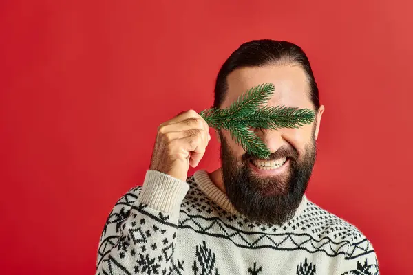 Joyful man in sweater covering eyes with branch of pine tree on red backdrop, Merry Christmas — Stock Photo