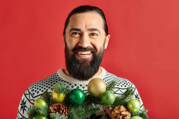 Joyeux homme barbu en pull d'hiver tenant décoré couronne de Noël avec des boules sur fond rouge — Photo de stock