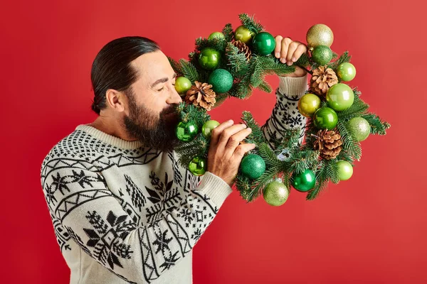 Heureux homme barbu en pull d'hiver tenant couronne décorée avec des boules sur fond rouge, Noël — Photo de stock