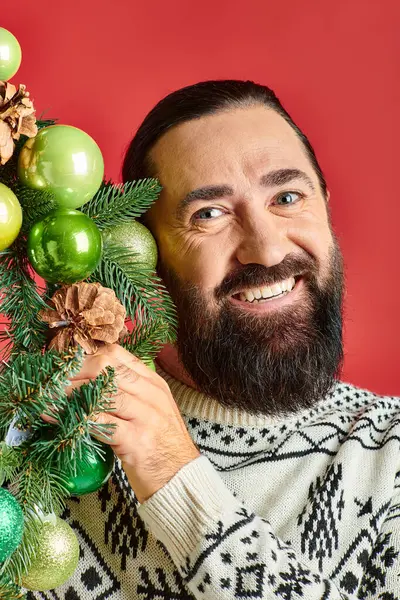 Веселый бородатый мужчина в зимнем свитере с украшенным рождественским венком с безделушками на красном фоне — стоковое фото