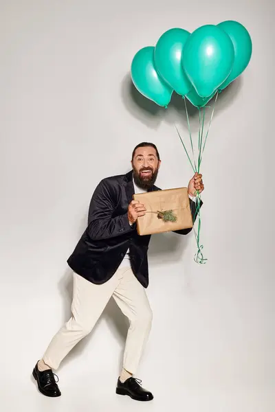 Happy bearded man in formal wear holding blue balloons and Christmas  present on grey backdrop — Stock Photo