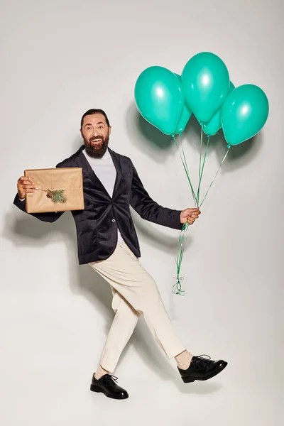 Excité homme barbu en tenue formelle tenant des ballons bleus et cadeau de Noël sur fond gris — Photo de stock