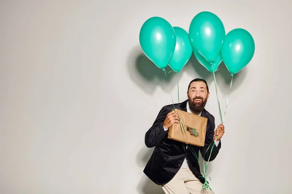Cheerful bearded man in formal wear holding blue balloons and Christmas  present on grey backdrop — Stock Photo