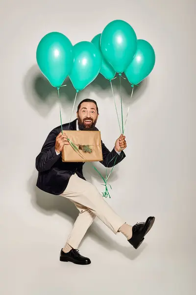 Drôle homme barbu en tenue formelle tenant des ballons bleus et cadeau de Noël sur fond gris — Photo de stock