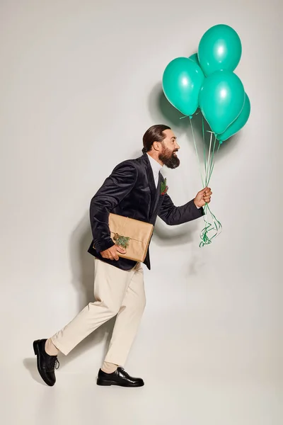 Homme barbu positif en tenue formelle tenant des ballons bleus et marchant avec cadeau de Noël sur gris — Photo de stock
