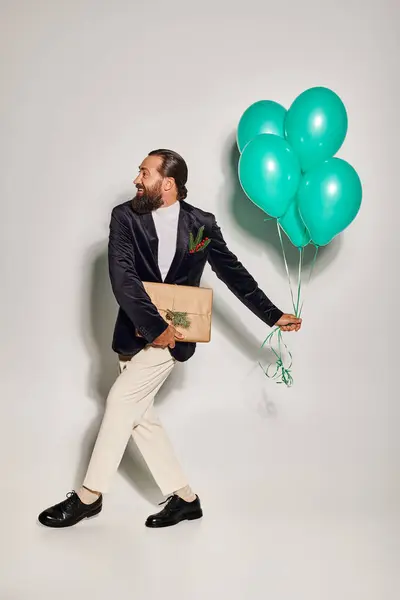 Happy bearded man in formal wear holding blue balloons and walking with Christmas present on grey — Stock Photo