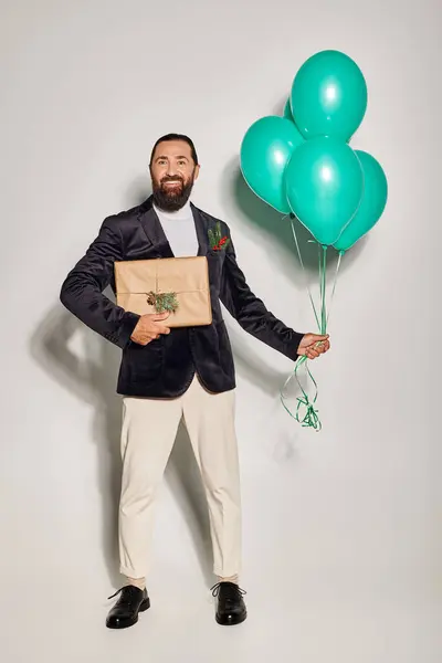 Heureux homme barbu en tenue formelle tenant des ballons bleus et debout avec cadeau de Noël sur gris — Photo de stock