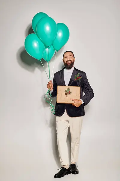 Smiling bearded man in formal attire holding blue balloons and Christmas  present on grey backdrop — Stock Photo
