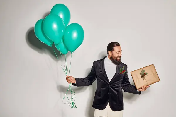 Cheerful bearded man in formal attire holding blue balloons and Christmas  present on grey backdrop — Stock Photo