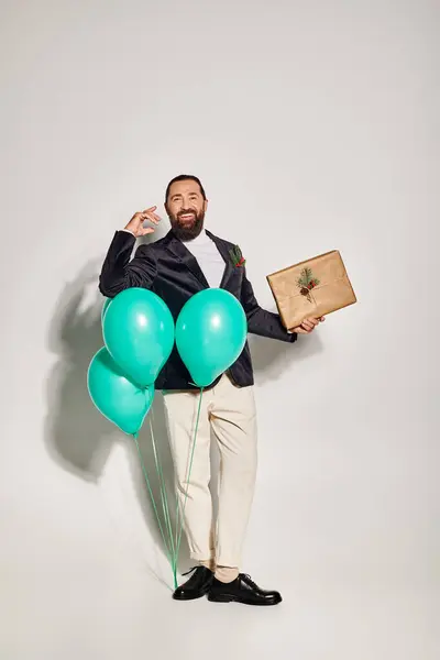 Homme barbu souriant en tenue formelle tenant cadeau de Noël près de ballons bleus sur fond gris — Photo de stock