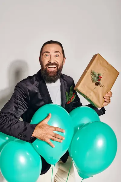 Joyeux homme barbu en tenue formelle tenant cadeau de Noël près de ballons bleus sur fond gris — Photo de stock