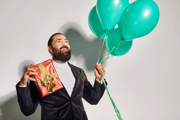 Joyful bearded man in formal wear holding Christmas present and blue balloons on grey backdrop — Stock Photo