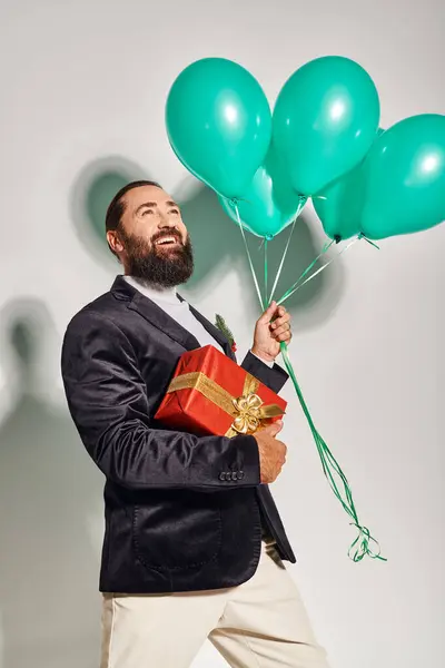 Fröhlicher bärtiger Mann in festlicher Kleidung mit Weihnachtsgeschenk und blauen Luftballons vor grauem Hintergrund — Stockfoto