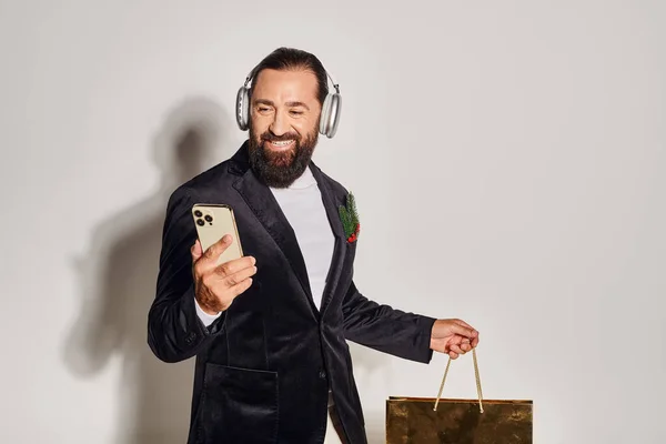 Happy bearded man in headphones and suit holding smartphone and shopping bag on grey backdrop — Stock Photo