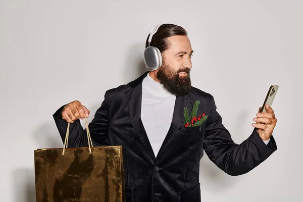 Homem barbudo feliz em fones de ouvido sem fio segurando smartphone e saco de compras no fundo cinza — Fotografia de Stock
