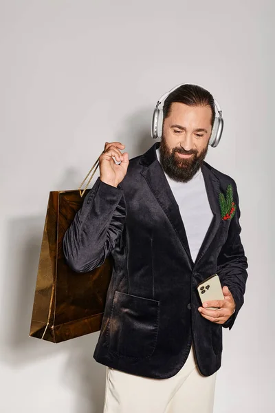 Homem barbudo alegre em fones de ouvido sem fio segurando smartphone e saco de compras no fundo cinza — Fotografia de Stock