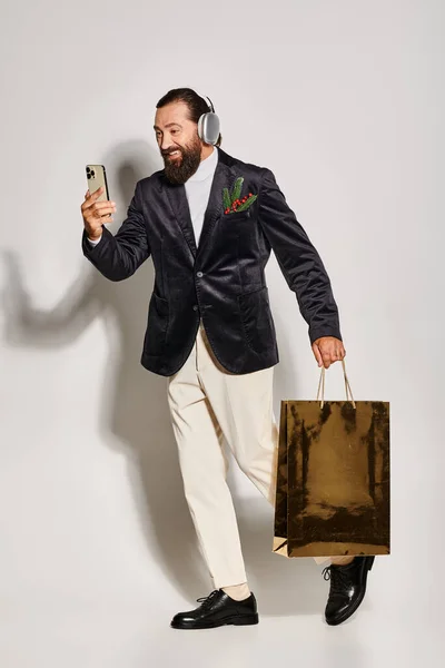 Happy bearded man in wireless headphones holding smartphone and gift bag on grey, video call — Stock Photo