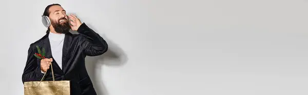 Homem alegre ouvindo música em fones de ouvido sem fio, segurando smartphone e saco do presente em cinza, banner — Fotografia de Stock