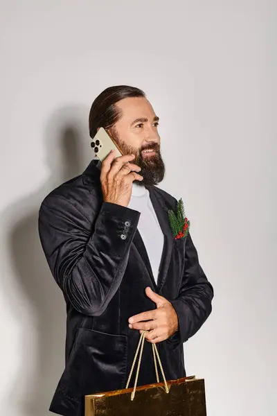 Щасливий бородатий чоловік у костюмі розмовляє на смартфоні і тримає різдвяний подарунковий пакет на сірому фоні — стокове фото