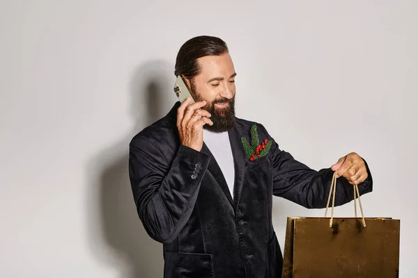 Happy bearded man in suit talking on smartphone and holding Christmas gift bag on grey backdrop — Stock Photo