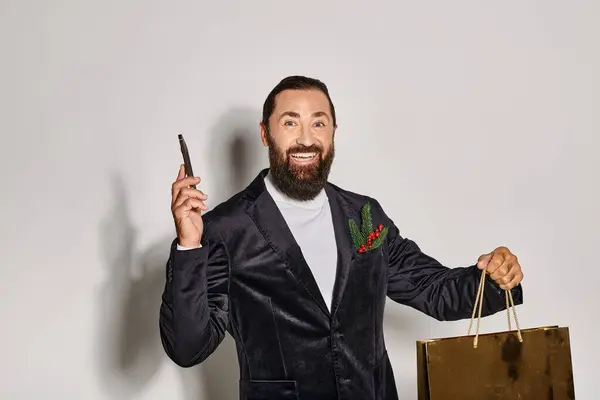 Счастливый бородатый человек в костюме держа смартфон и рождественский подарочный пакет на сером фоне, покупки — стоковое фото