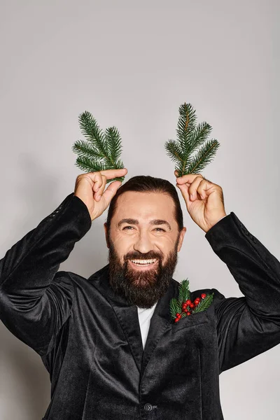 Hombre barbudo feliz en traje sosteniendo dos ramas de abeto cerca de la cabeza y sonriendo en el fondo gris - foto de stock