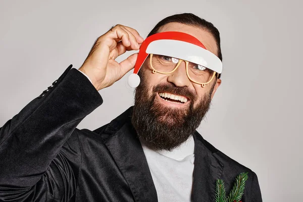 Happy bearded man in festive glasses with santa hat posing in suit and smiling on grey backdrop — Stock Photo