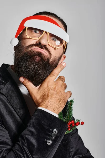 Bärtiger Mann mit festlicher Brille und Weihnachtsmütze posiert im Anzug und schmollenden Lippen vor grauem Hintergrund — Stockfoto
