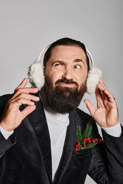 Емоційний бородатий чоловік у костюмі з різдвяною ялинкою в кишені, що носить вушні муфти на сірому тлі — стокове фото