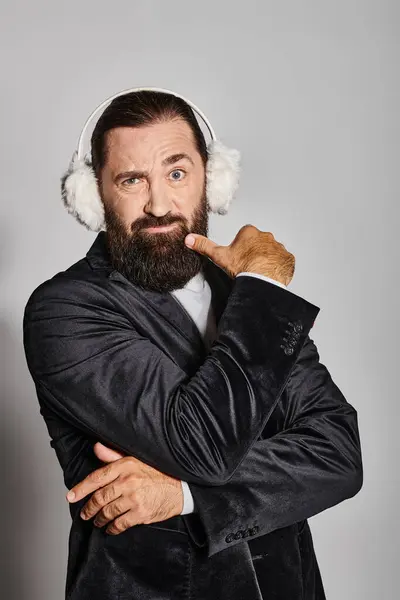 Dudoso hombre barbudo en traje con ramas de abeto de Navidad y orejeras moviendo las cejas en gris - foto de stock