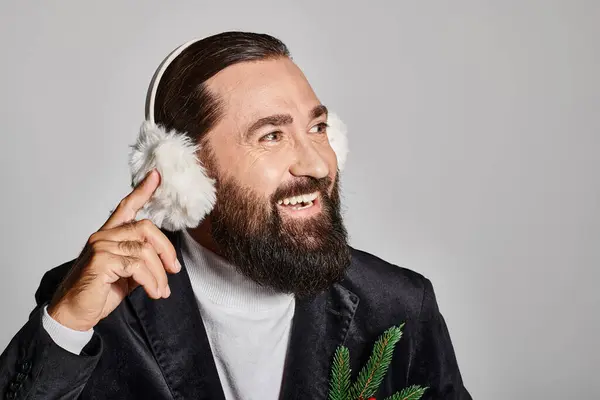 Cheerful bearded man in suit with Christmas spruce branch and red berries wearing ear muffs on grey — Stock Photo