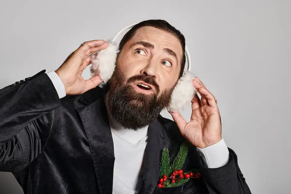Zerstreuter bärtiger Mann im Anzug mit weihnachtlichen Fichtenzweigen, die Ohrenschützer vor grauem Hintergrund berühren — Stockfoto