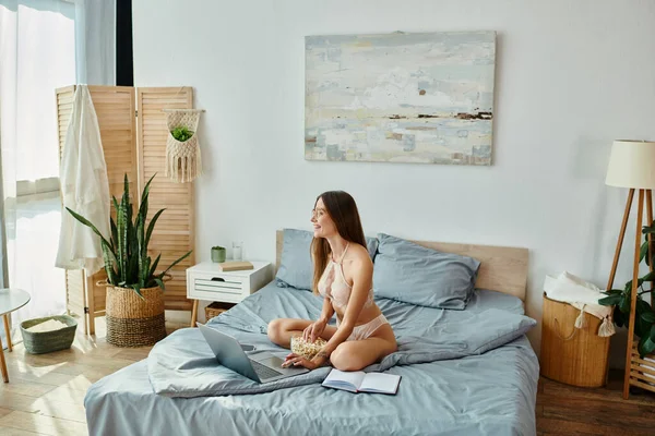 Mulher sexy alegre em lingerie bege com óculos sentados na cama com pipoca e trabalhando remotamente — Fotografia de Stock