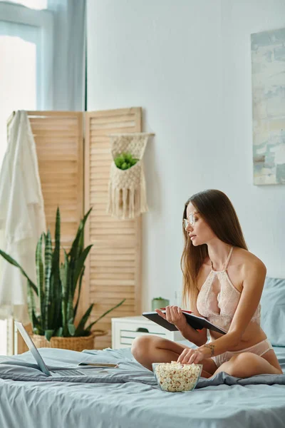Jeune femme réfléchie en lingerie beige avec des lunettes manger du pop-corn et travailler à distance sur un ordinateur portable — Photo de stock