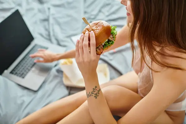 Vista cortada de jovem com cabelo comprido comendo delicioso hambúrguer enquanto trabalhava em seu laptop — Fotografia de Stock
