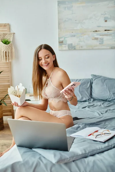 Mujer alegre en lencería sentada en la cama sosteniendo fideos y teléfono y sonriendo a su cámara portátil - foto de stock