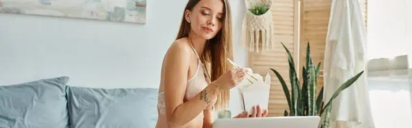 Mujer pensativa en lencería comiendo fideos y mirando a la cámara del ordenador portátil mientras trabaja duro, pancarta - foto de stock