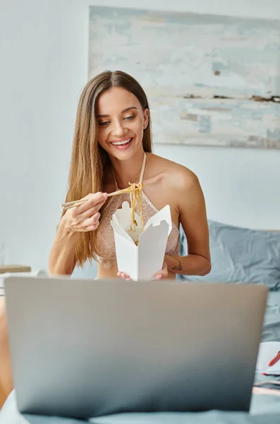Alegre joven mujer comiendo fideos con palillos y mirando a su computadora portátil mientras trabaja duro - foto de stock